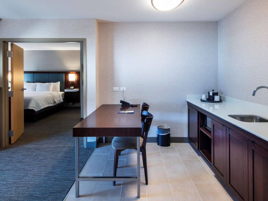A compact hotel room with a separate dining area, a bed, a bedside table, a lamp, and a small trash bin.