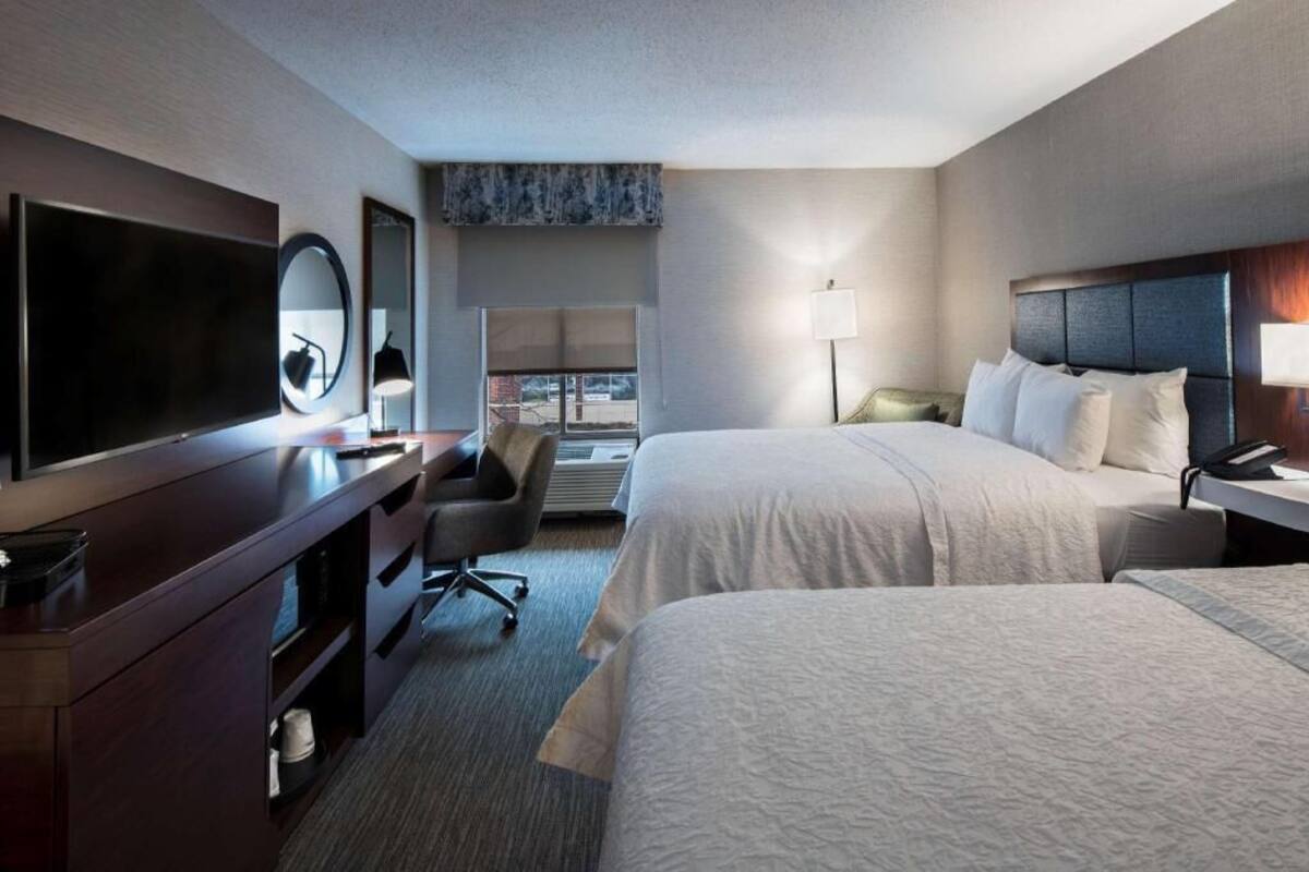 A hotel room with a flat-screen TV, a desk with a chair, two beds, a window, and a lamp.