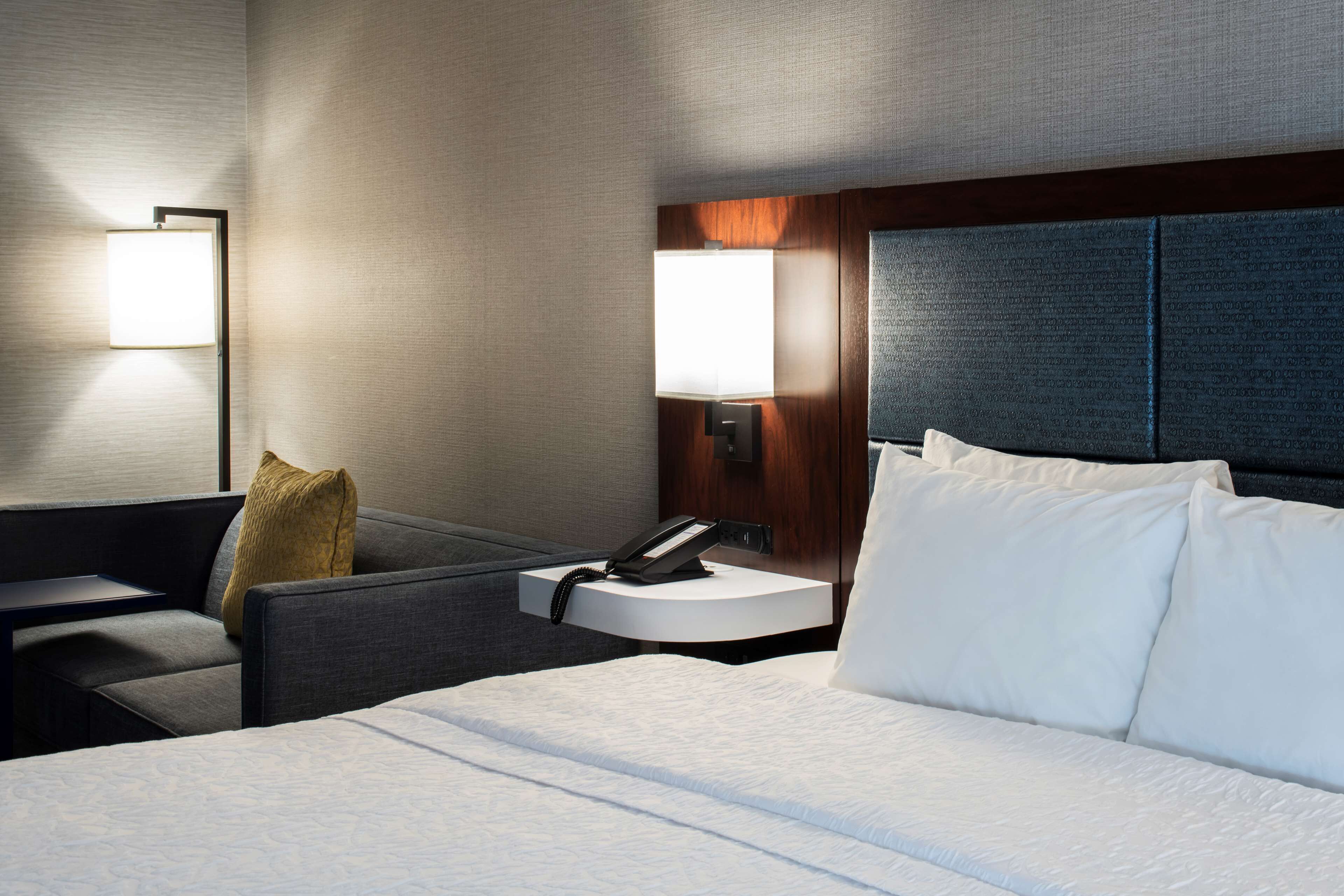 Une chambre d’hôtel comprenant un lit, un bureau, une chaise, une table de chevet avec un téléphone et deux lampes fixées au mur.