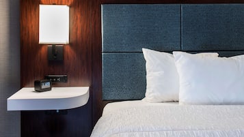 Ein Hotelzimmer mit einem hölzernen Kopfteil, einem Nachttisch mit digitaler Uhr und einem weißen Bett mit Kissen.