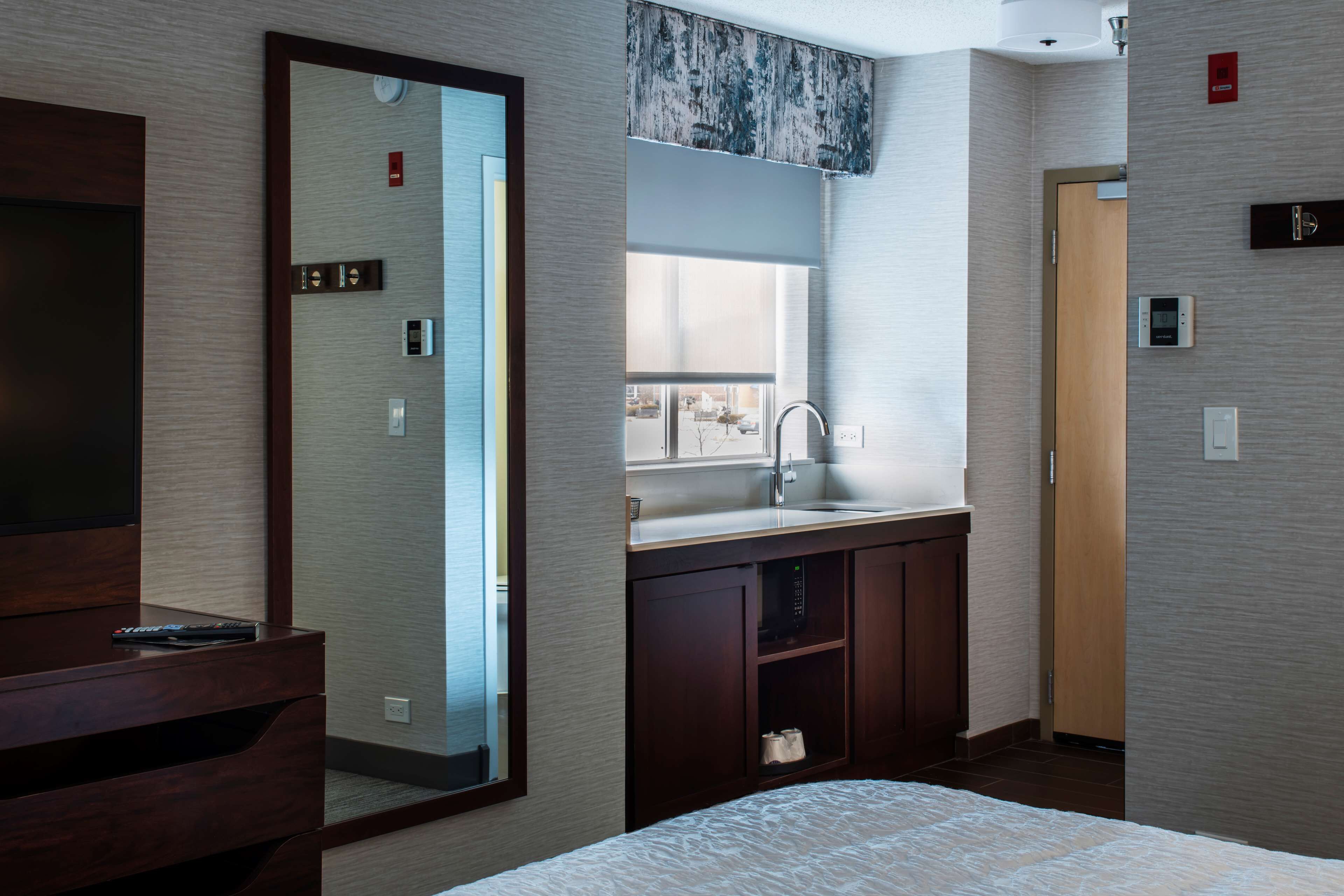 Habitación de hotel con un espejo grande, un tocador de madera con lavamanos, una cama y vistas al exterior a través de una ventana.