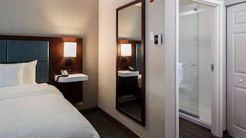 A hotel room with a bed, a bedside table with a lamp, a mirror, a bathroom with a sink, and a shower.