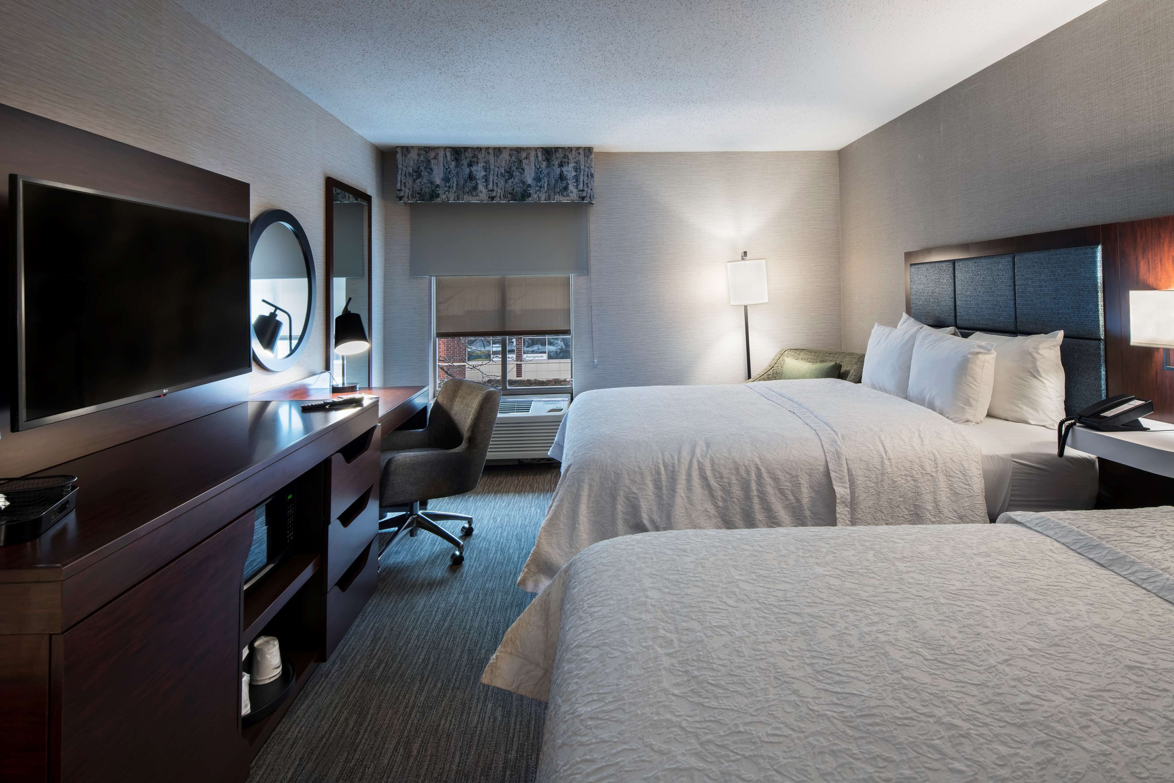 A hotel room with two beds, a TV, a desk with a chair, a lamp, and a large window.