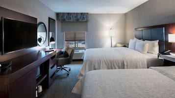 A hotel room with two beds, a TV, a desk with a chair, a lamp, and a large window.