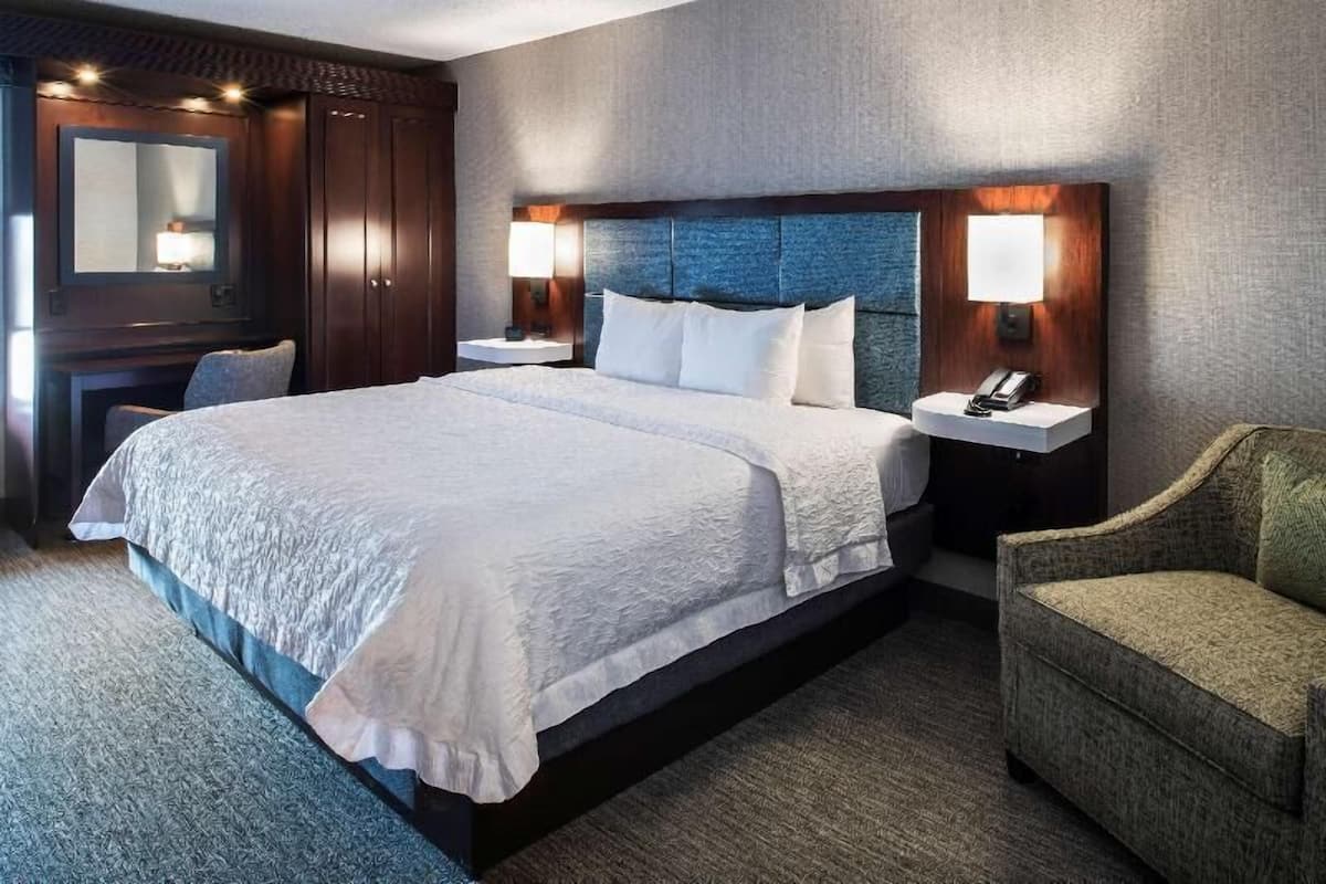 A hotel room with a large bed, two bedside tables, a chair, a mirror, and a wall-mounted lamp.