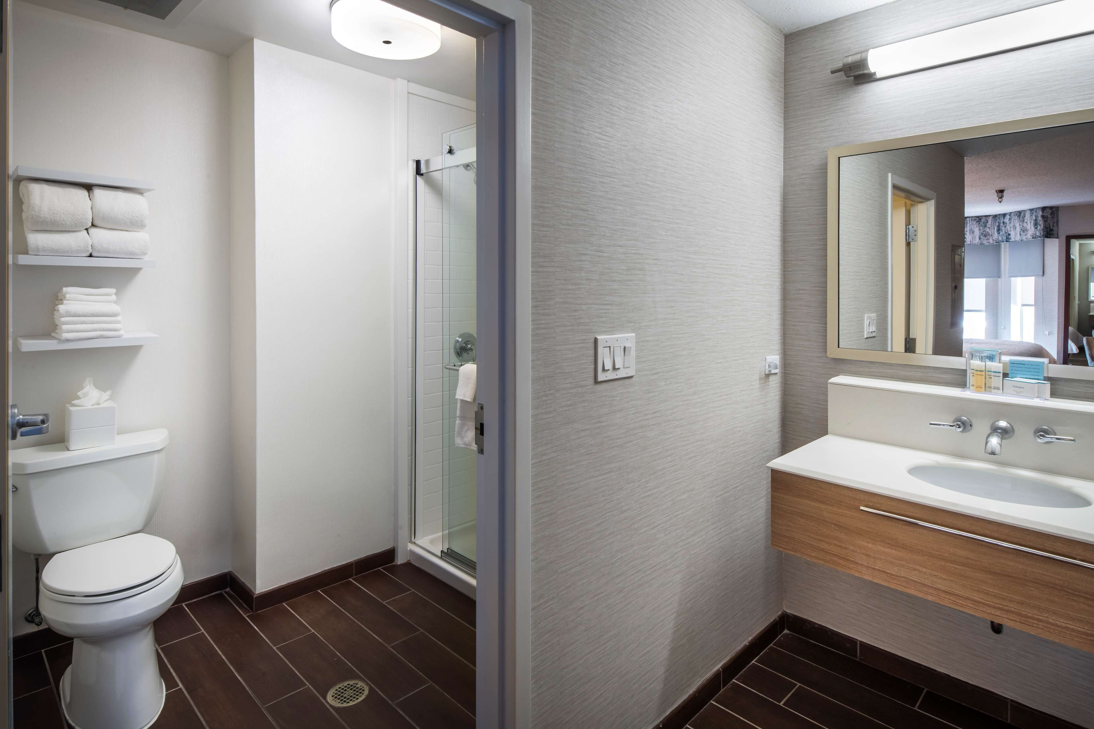 A modern bathroom with a toilet, a glass shower enclosure, a sink with a mirror, and shelves with towels.