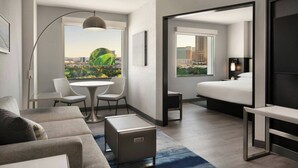A modern hotel room with a large bed, a seating area with a table and chairs, a small side table, and a view of the cityscape through large windows. - Las Vegas Marriott (Las Vegas)