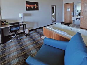 A modern hotel room with a large bathtub, a blue sofa, a desk with a chair, a mirror, and a picture of a bridge on the wall. - AmericInn by Wyndham Saint Ignace Lakefront (St Ignace)