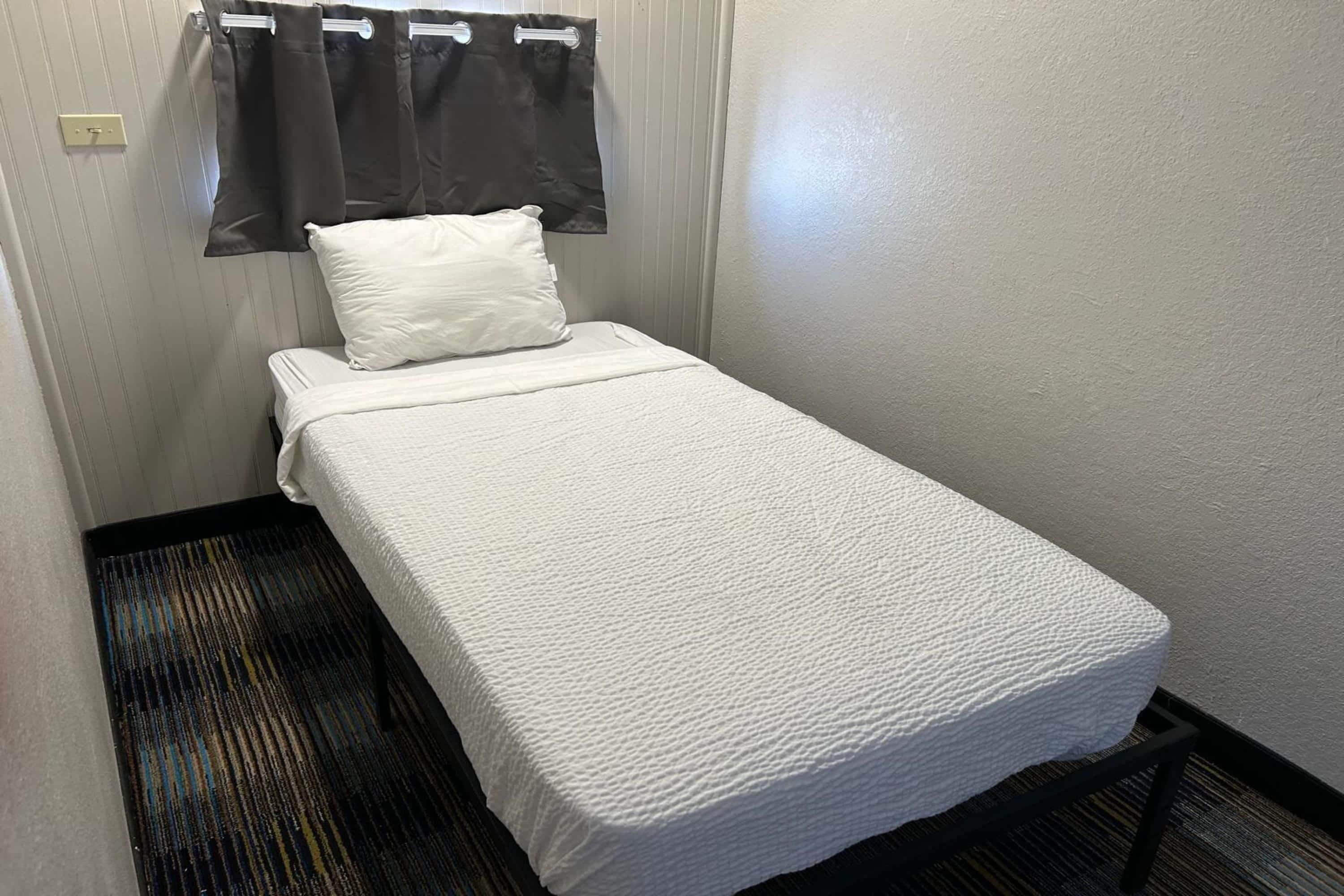 Une petite chambre avec un lit simple, recouverte d’une courtepointe blanche, un oreiller et un rideau de couleur sombre.