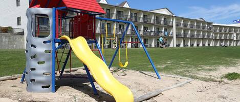 A playground with a yellow slide and a red-roofed playhouse, set against a backdrop of a multi-story hotel with balconies.