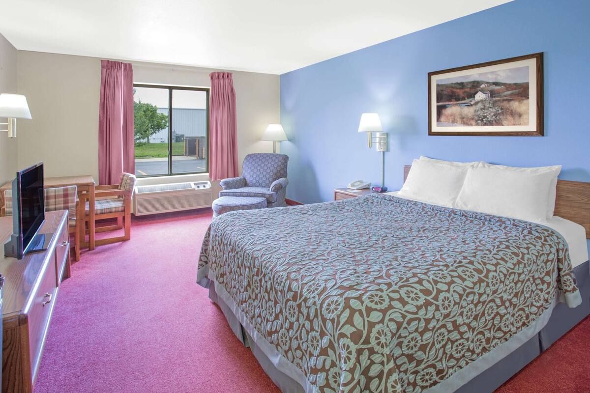 A hotel room with a bed, a desk with a computer, a chair, a window with curtains, and a painting on the wall.
