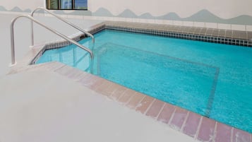 Una piscina con borde de azulejos, una escalera metálica y vista a un estacionamiento a través de una ventana.
