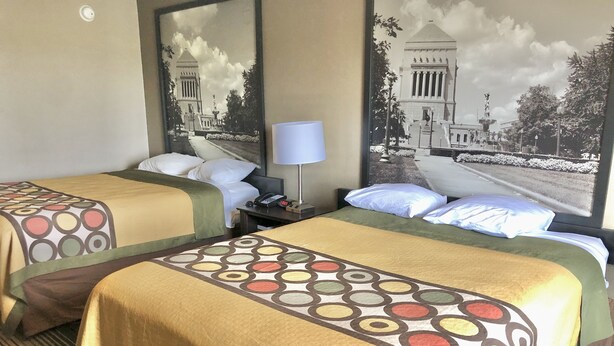 A hotel room with two beds, each with a patterned bedspread, a bedside table with a lamp, and a large mirror reflecting a building.