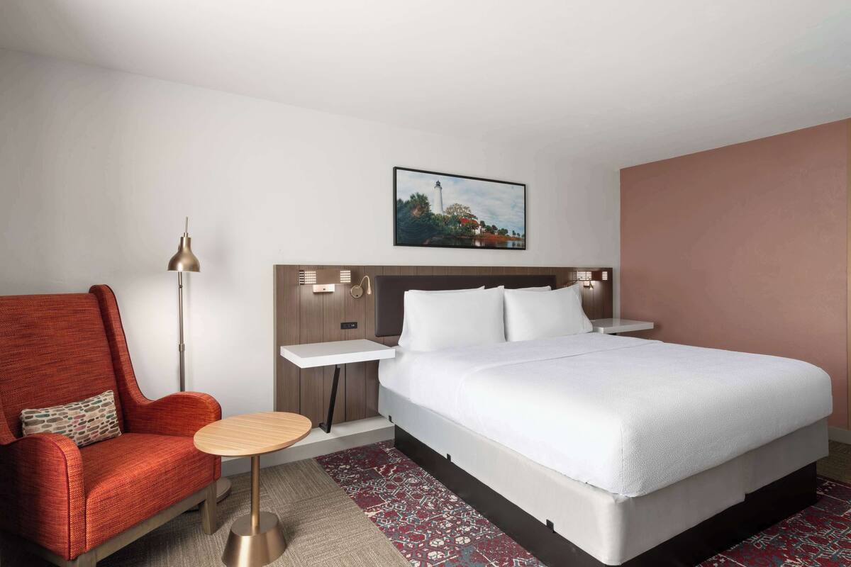 A hotel room with a large bed, a red armchair, a small round table, a floor lamp, and a painting on the wall.