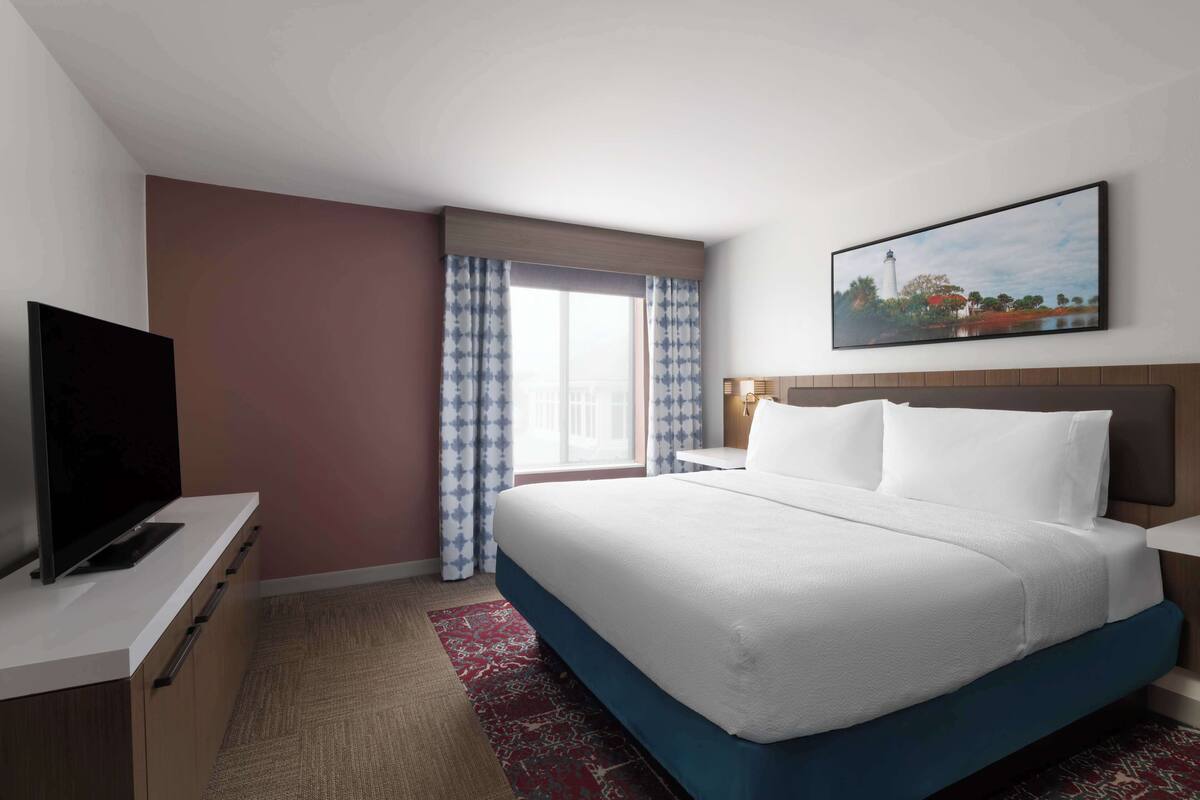 A hotel room with a bed, a flat-screen TV, a painting, and a window with curtains.