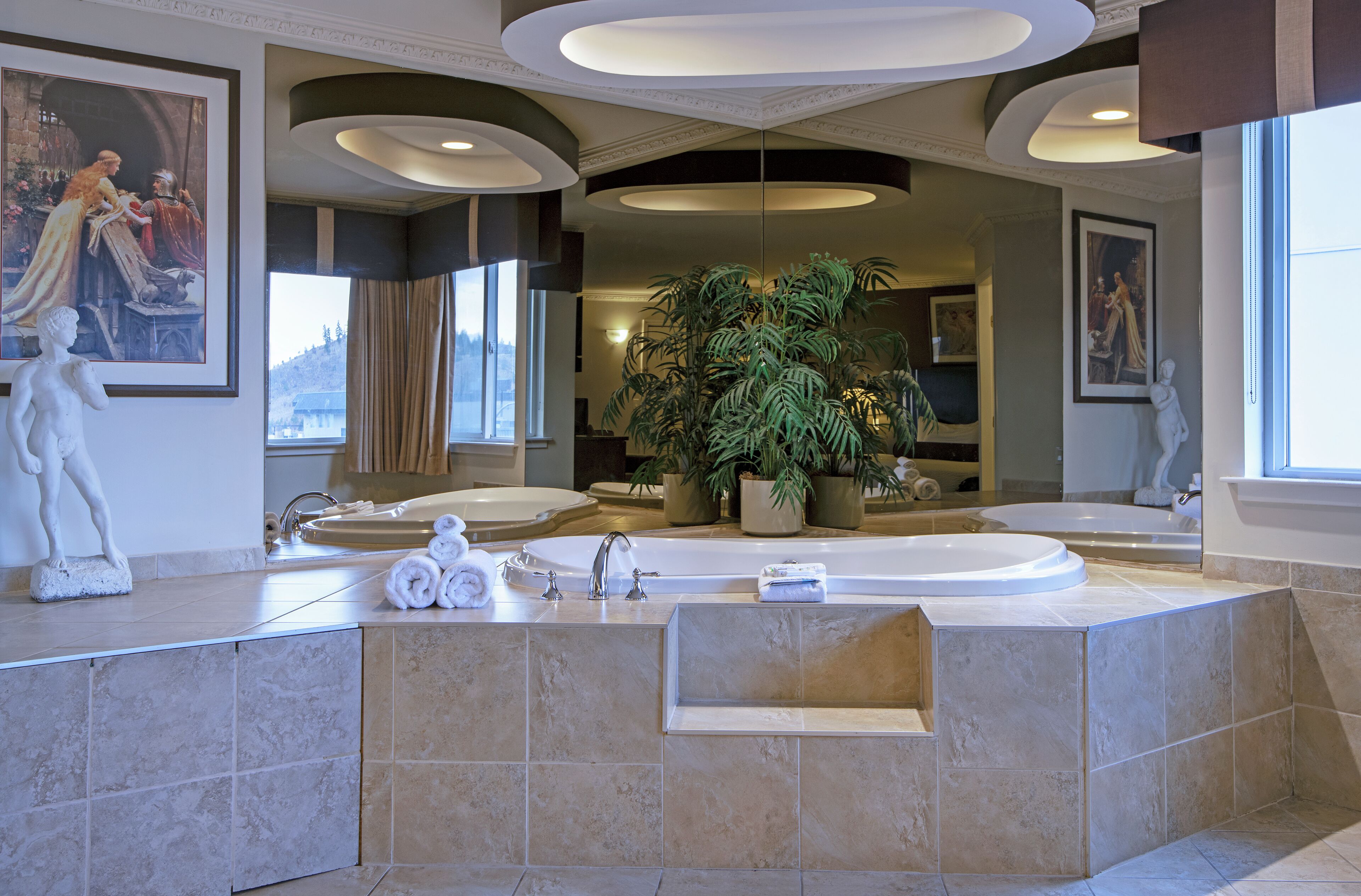 A luxurious bathroom with a large bathtub, a sink, and a mirror. There are decorative statues and a large potted plant.