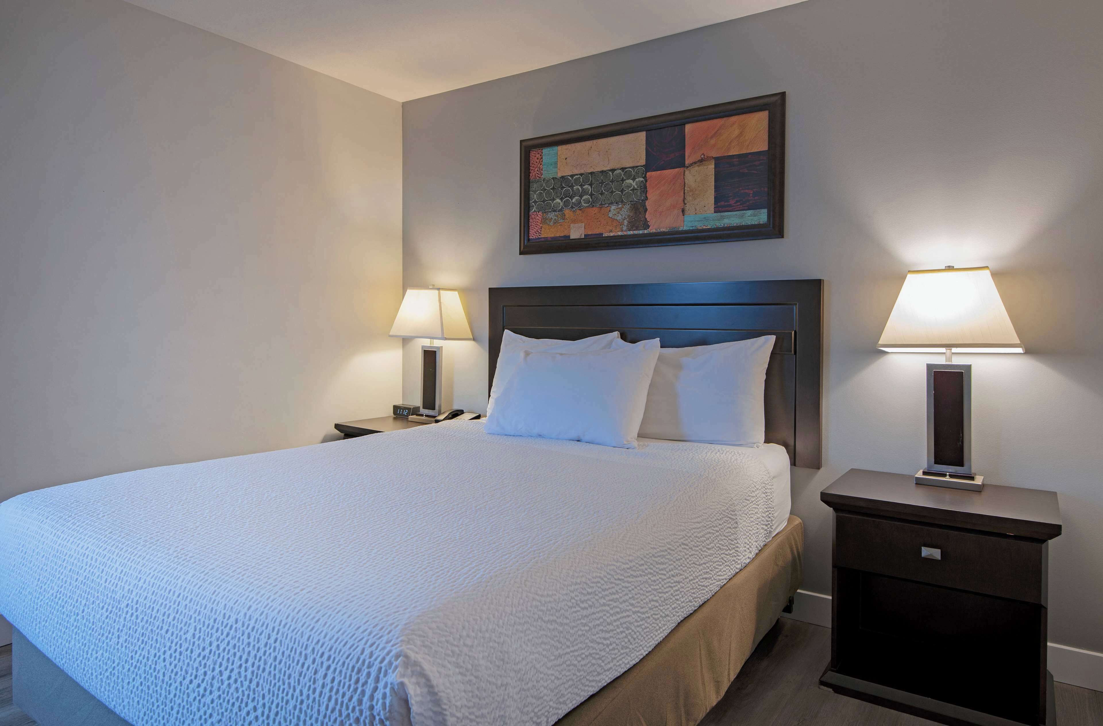 A neatly made bed with white linens, two bedside tables with lamps, and a colorful abstract painting on the wall.