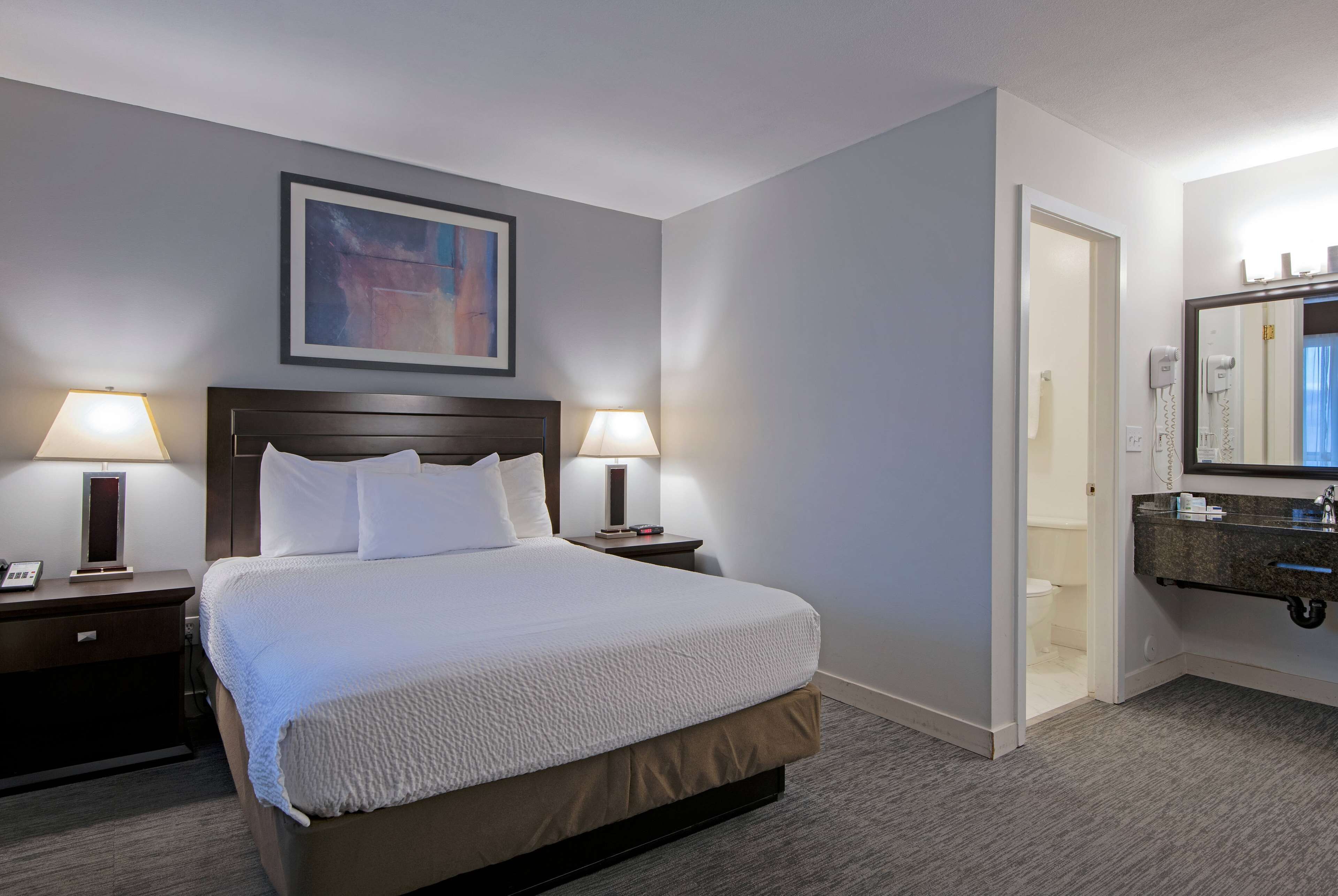 A hotel room with a large bed, two bedside lamps, a framed artwork on the wall, and a bathroom with a mirror and sink.