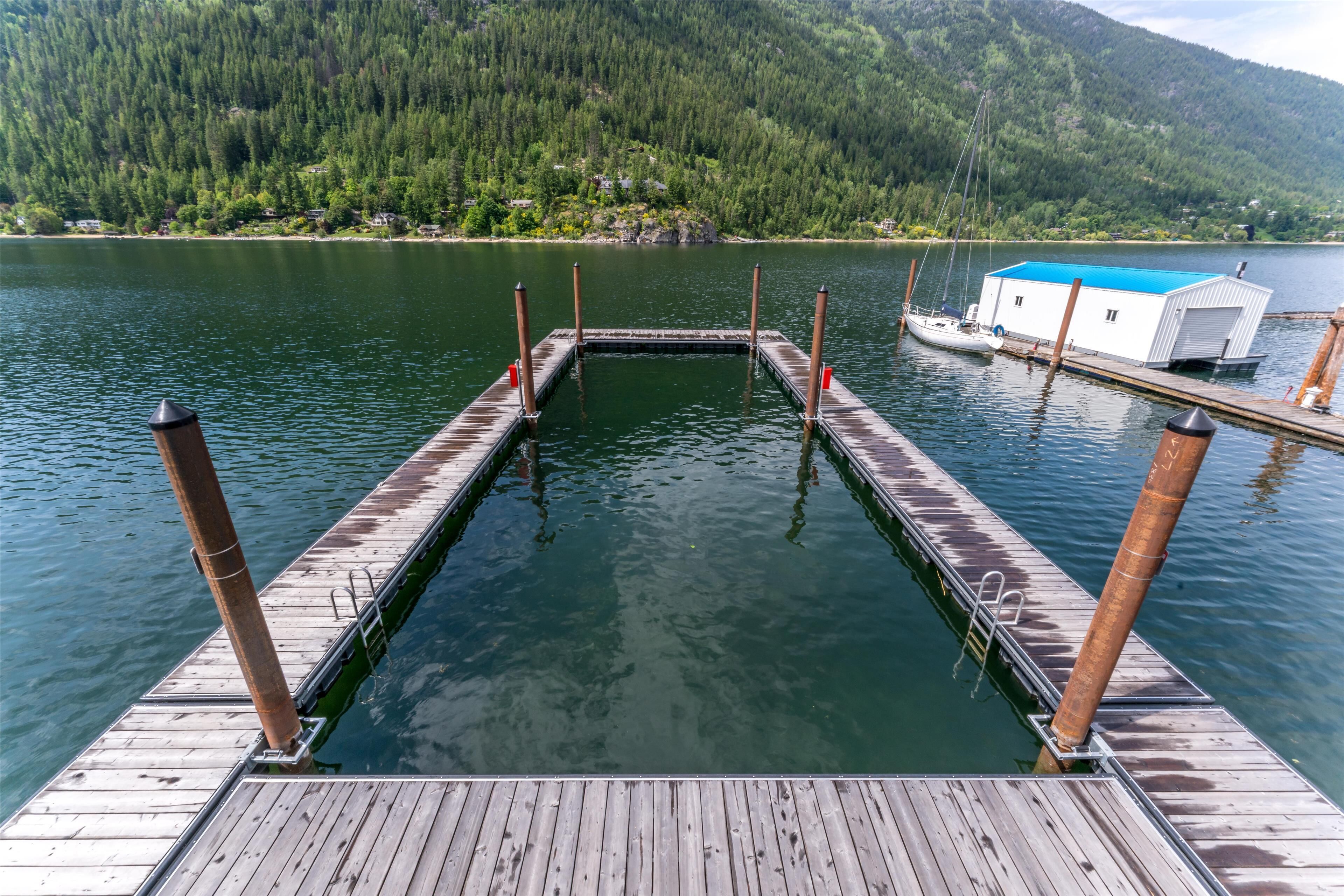 Um cais de madeira que se estende por um lago calmo, rodeado por uma floresta densa e um pequeno edifício com telhado azul.