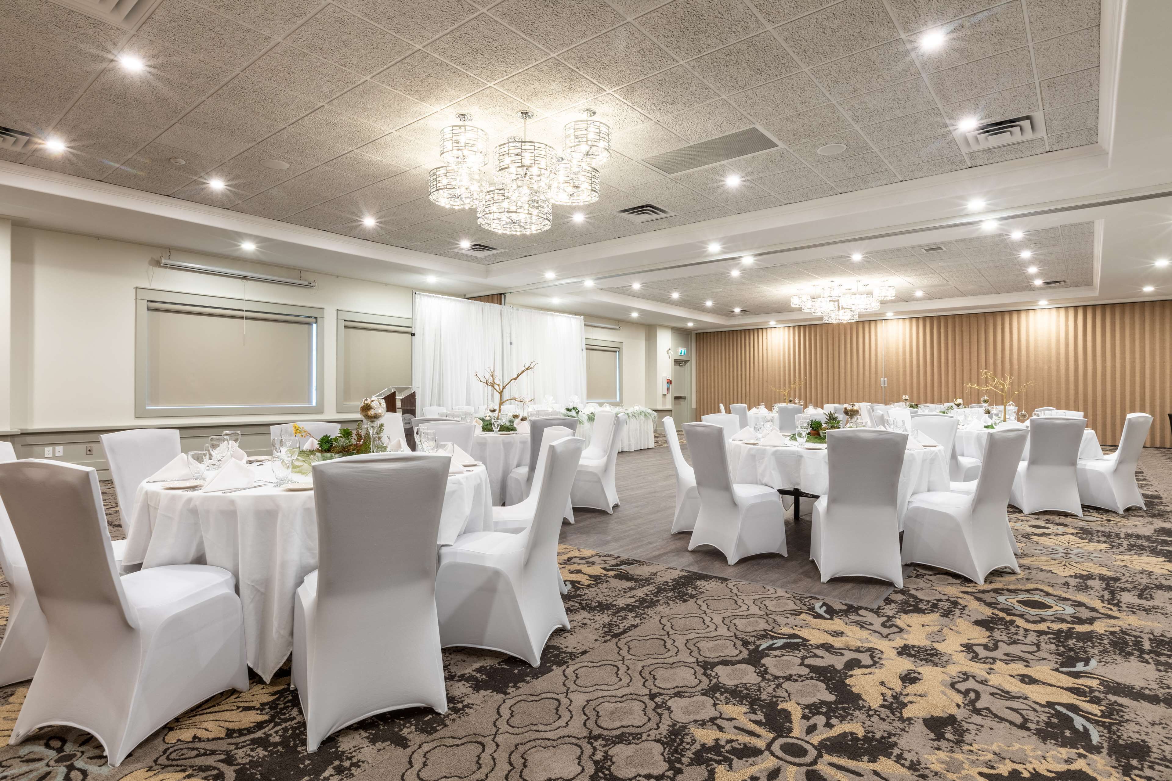 Un salón de banquetes con mesas redondas, sillas blancas, candelabros y suelo alfombrado.