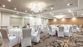 Un salón de banquetes con mesas redondas, sillas blancas, candelabros y suelo alfombrado.