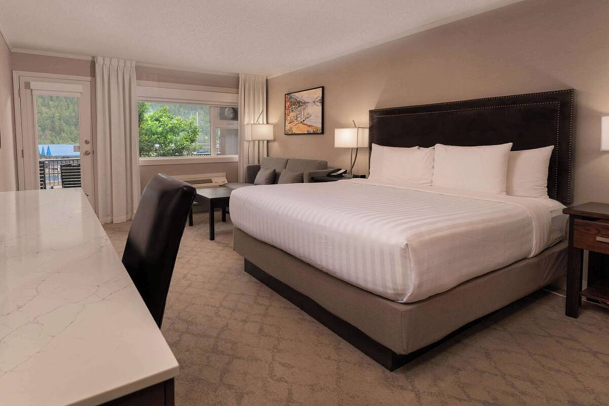 Quarto de hotel com uma cama grande, uma escrivaninha, uma cadeira, uma mesa de cabeceira e uma janela com cortinas.