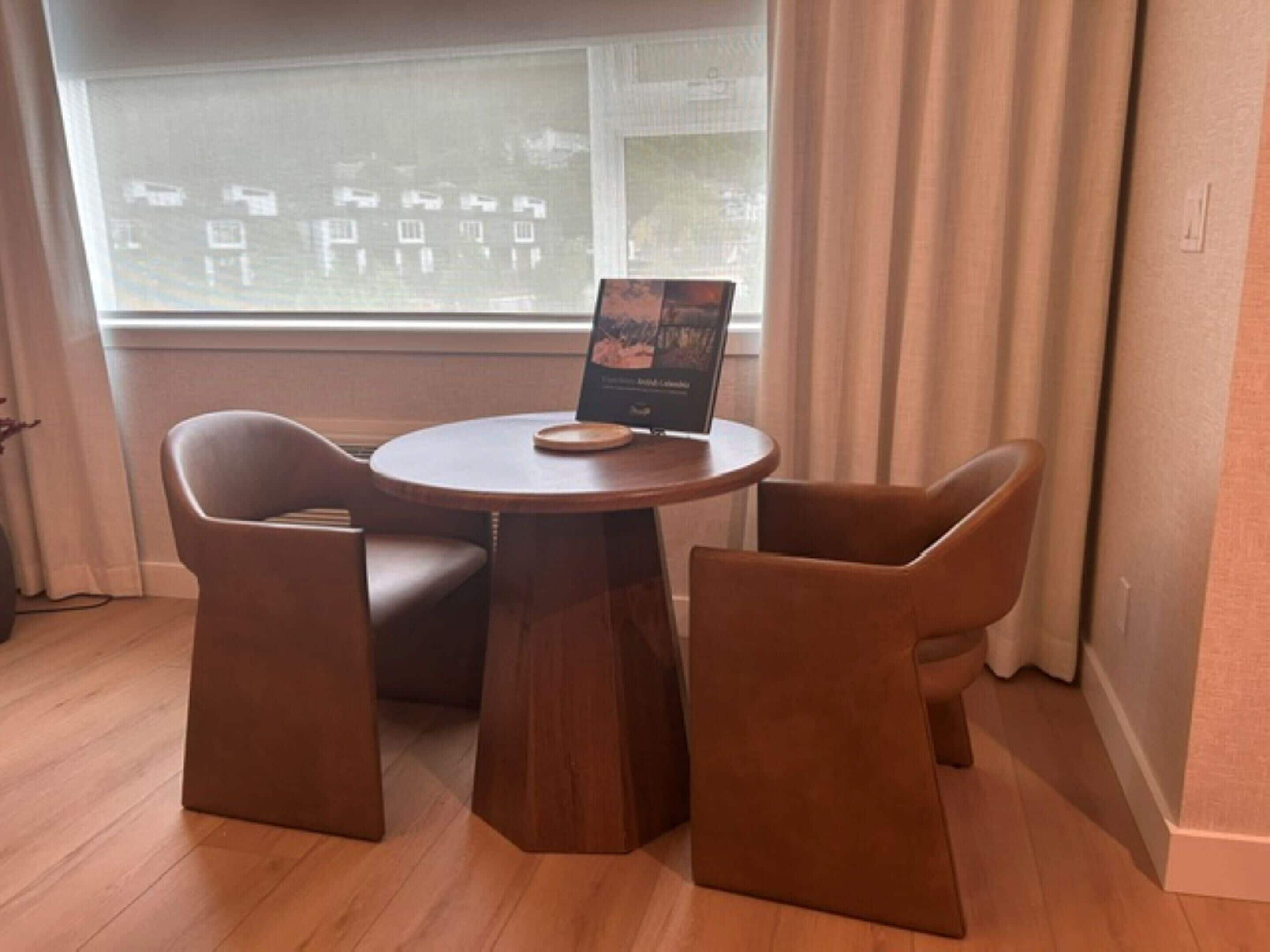 Una habitación con una mesa redonda de madera, dos sillas marrones, una ventana con cortinas y un cuadro en la mesa.