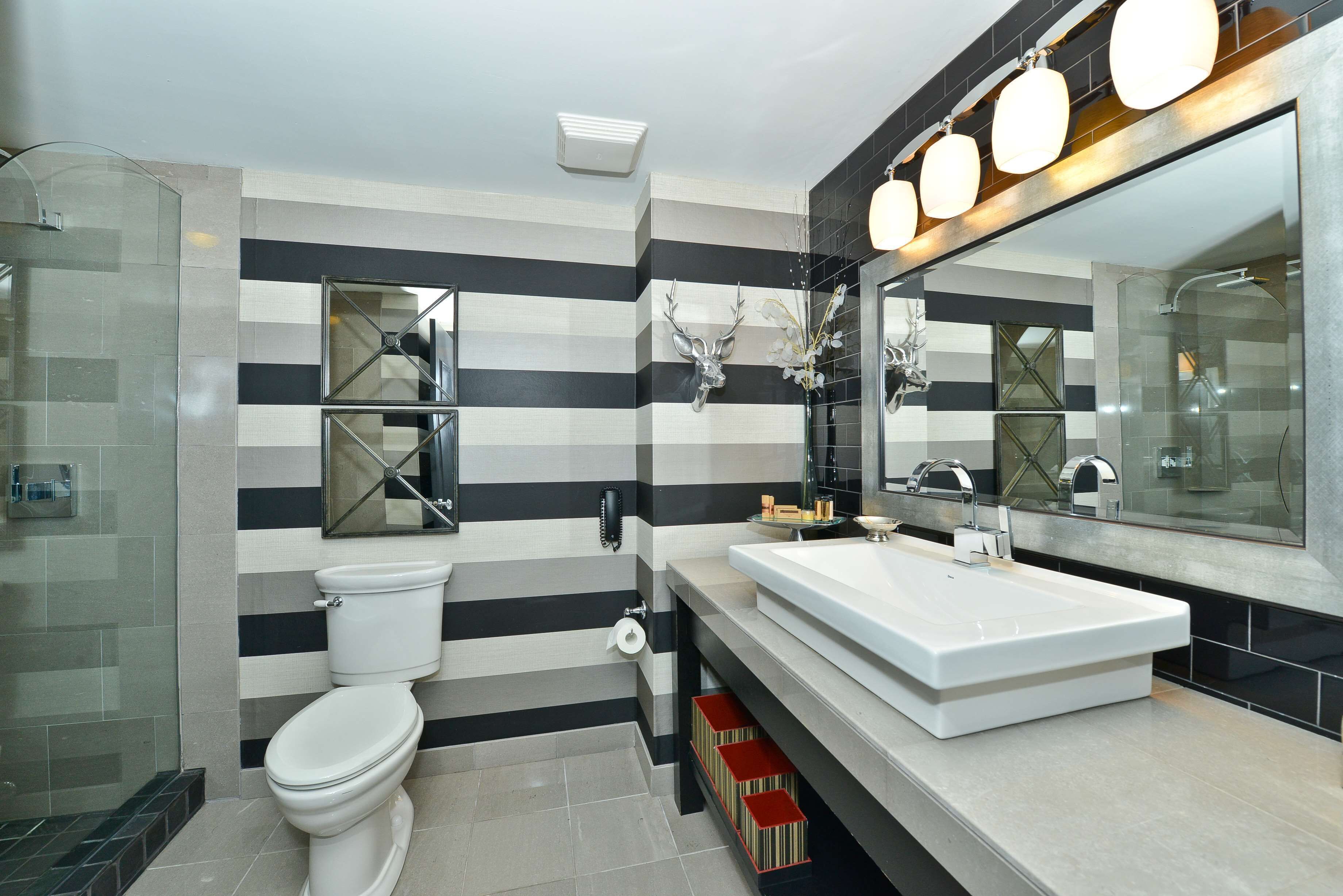 Une salle de bain moderne avec un mur à rayures, un grand miroir, un lavabo blanc et une cabine de douche en verre.