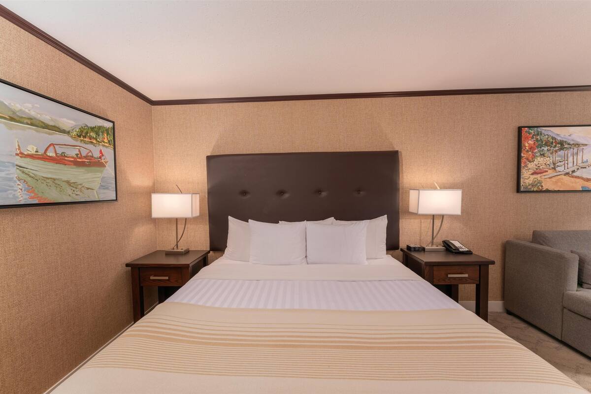 Quarto de hotel com uma cama grande, duas mesas de cabeceira, uma cadeira e duas luminárias fixadas na parede.