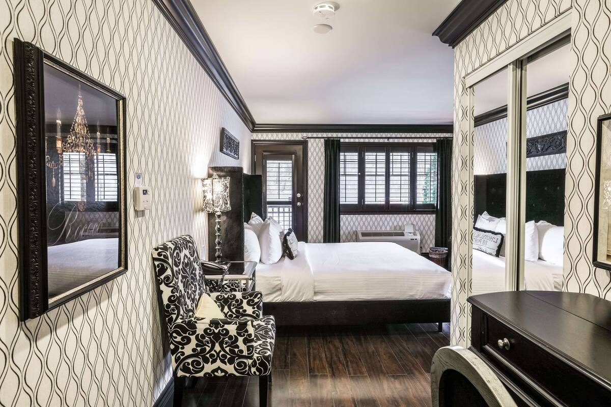 Quarto de hotel com uma cama grande, uma cadeira, uma mesa de escritório e uma cômoda. Há duas janelas com cortinas, um candelabro e um espelho na parede.