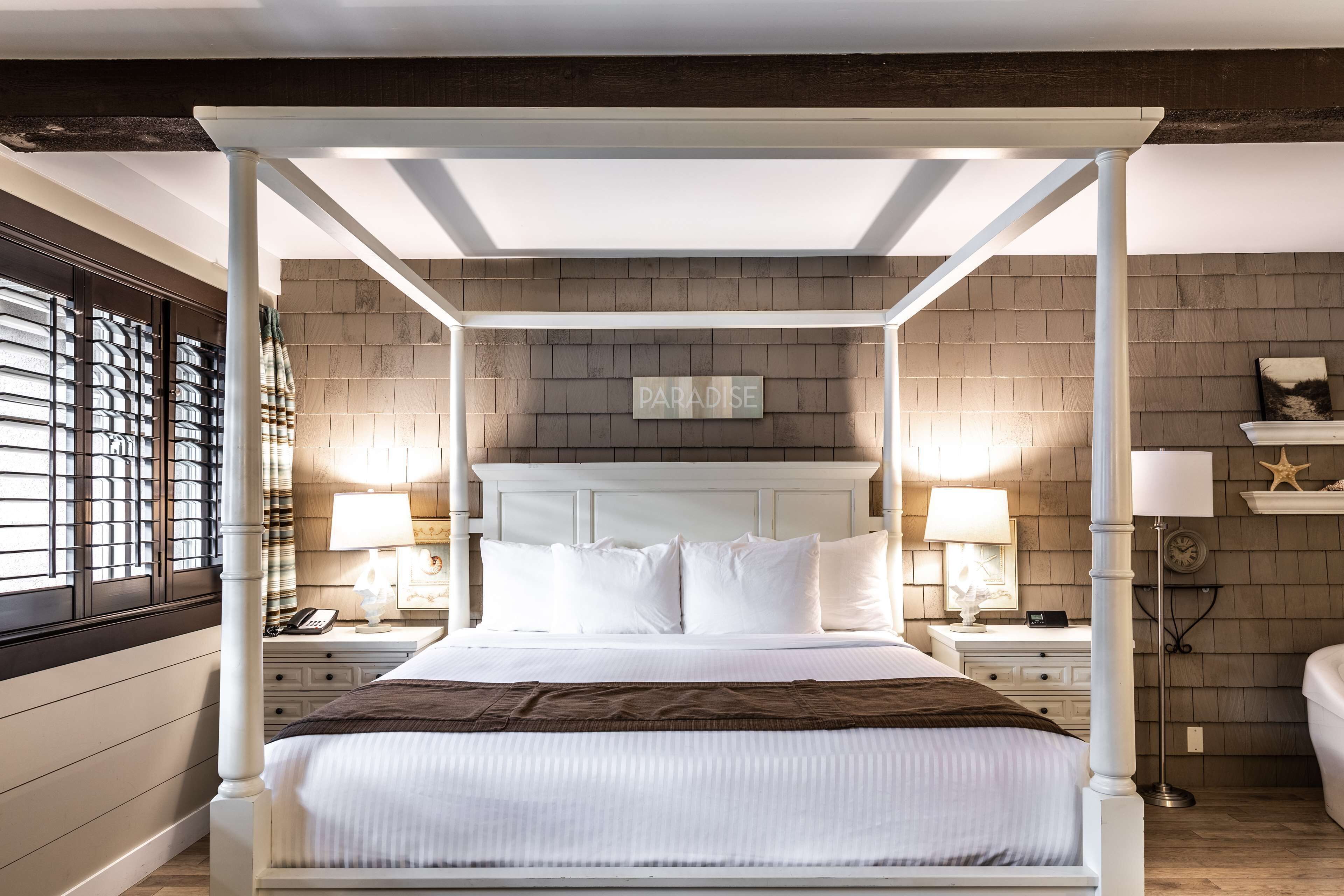 A four-poster bed with white bedding, a wooden headboard, and a brick-patterned wall behind it.