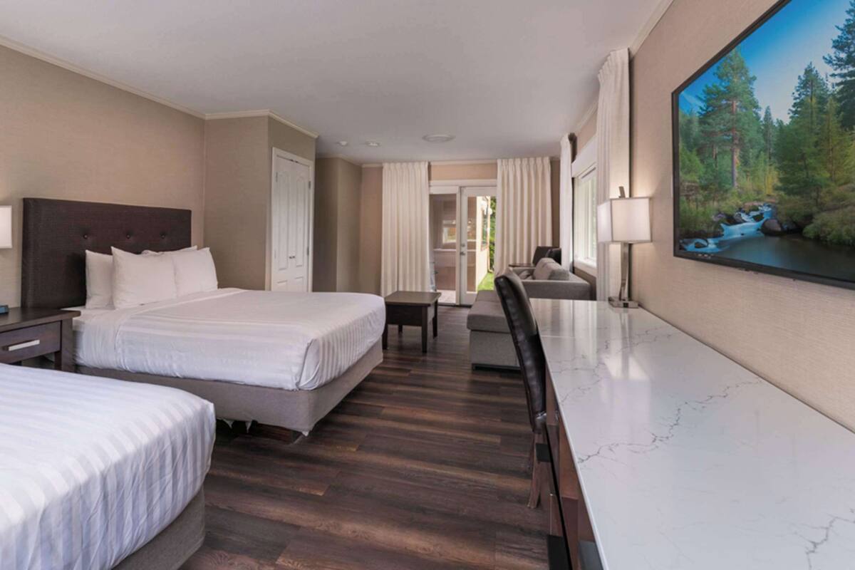 Quarto de hotel com duas camas, uma mesa de jantar e uma janela grande com vista para uma floresta.