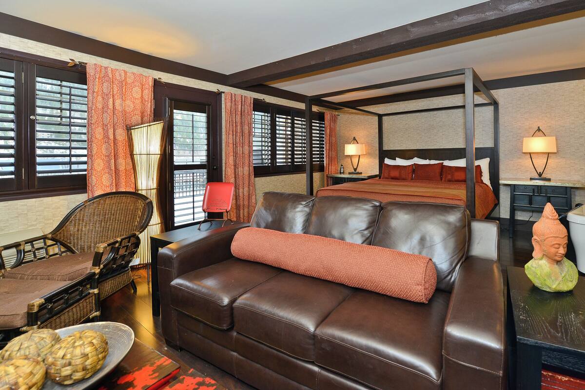 Quarto de hotel com sofá de couro marrom, cama com roupa de cama vermelha, mesa pequena com estátua de Buda e uma cadeira.