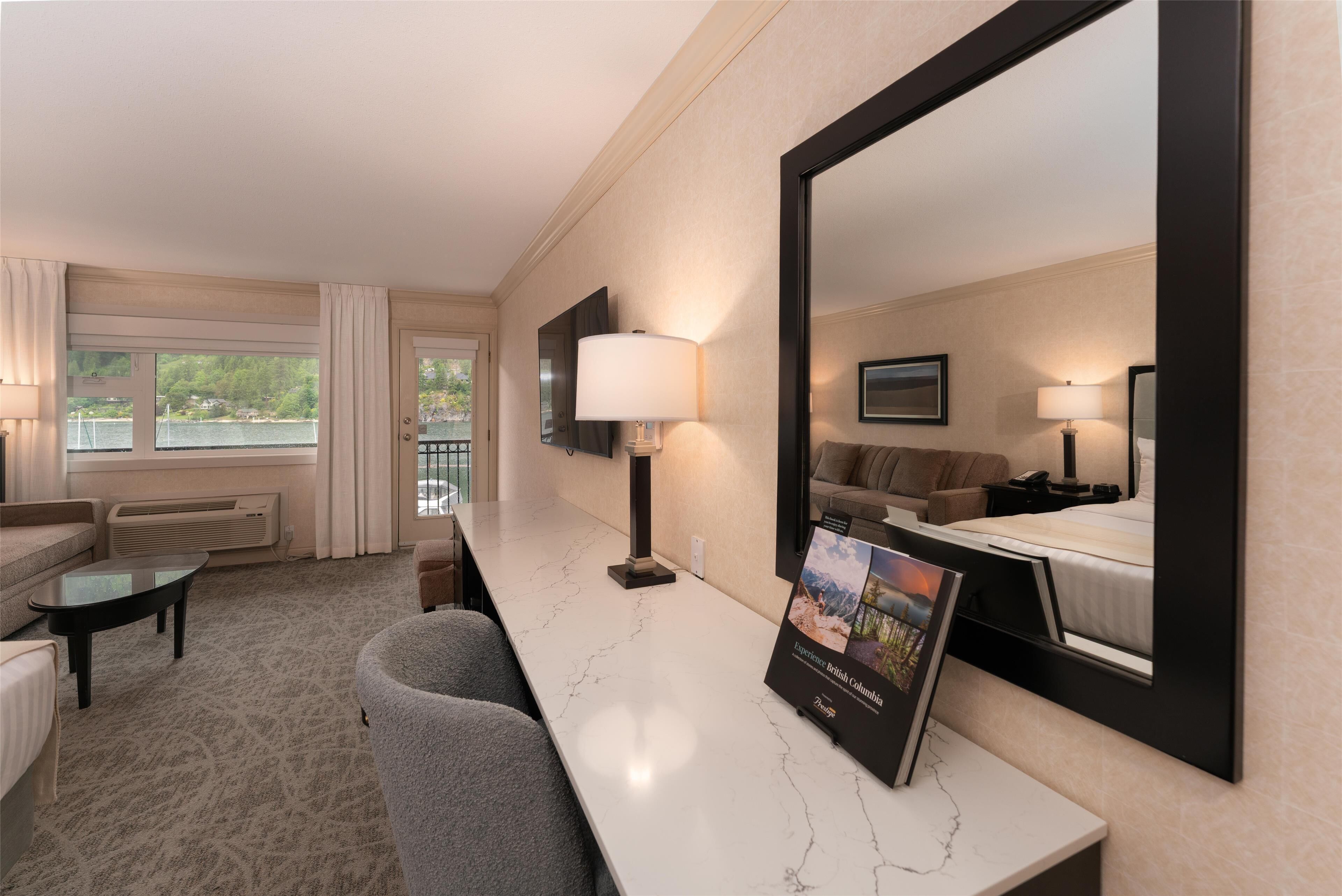 A hotel room with a large bed, a sitting area with a sofa and armchairs, a marble-topped desk, a large mirror, and a view of a body of water through the windows.