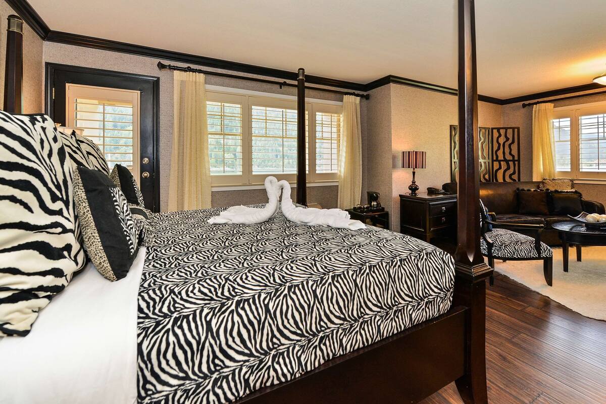 Uma cama com dossel com roupa de cama estampada em zebra, um espaço de estar com sofá e poltrona, e uma mesa de jantar com uma tigela de frutas.