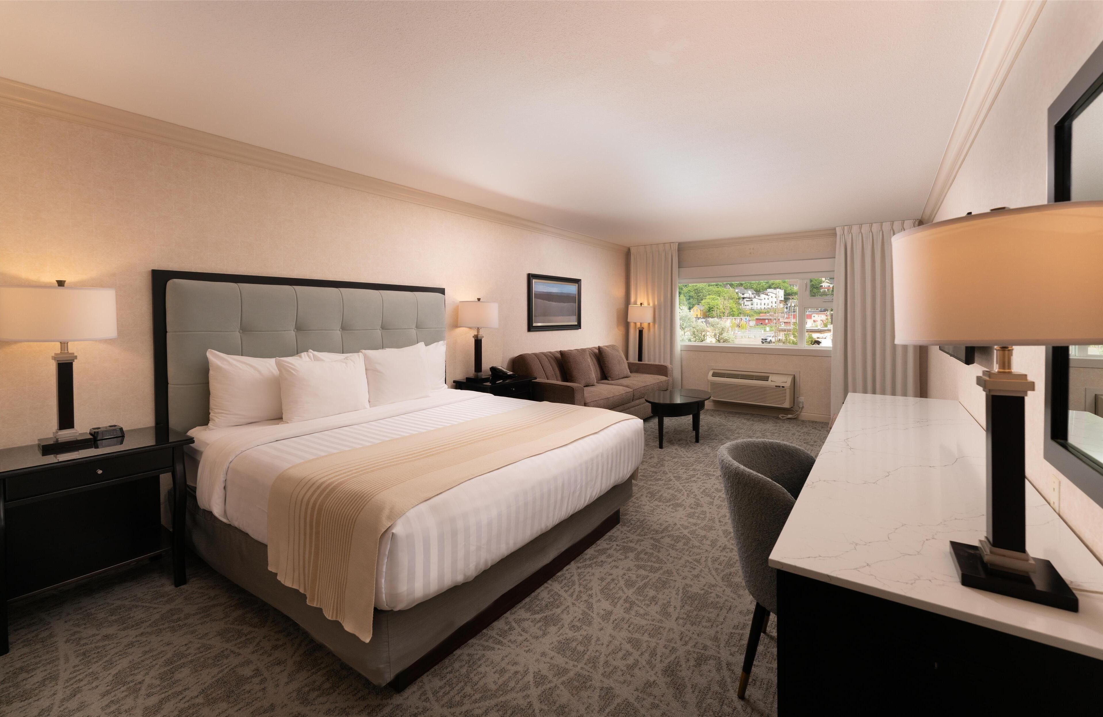 A hotel room with a large bed, a desk, a chair, a sofa, and a window with a view of the city.