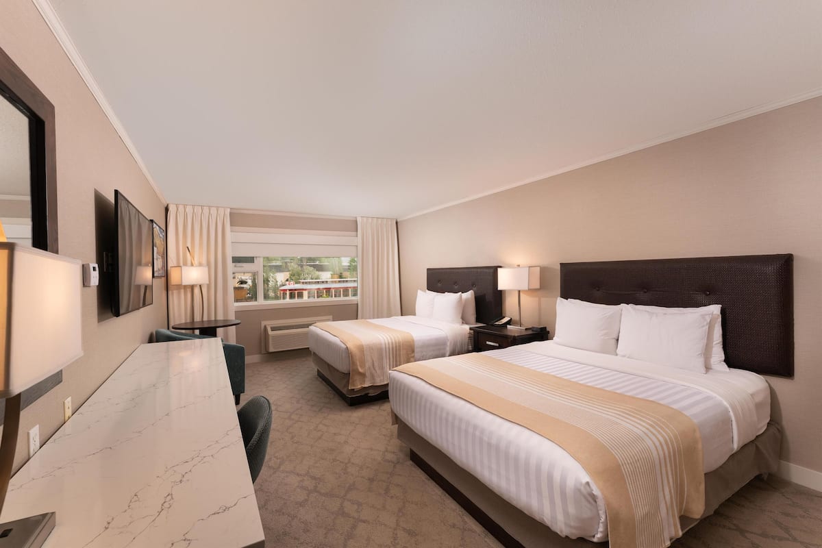 Quarto de hotel com duas camas, uma escrivaninha, uma cadeira, uma televisão e uma janela com cortinas.