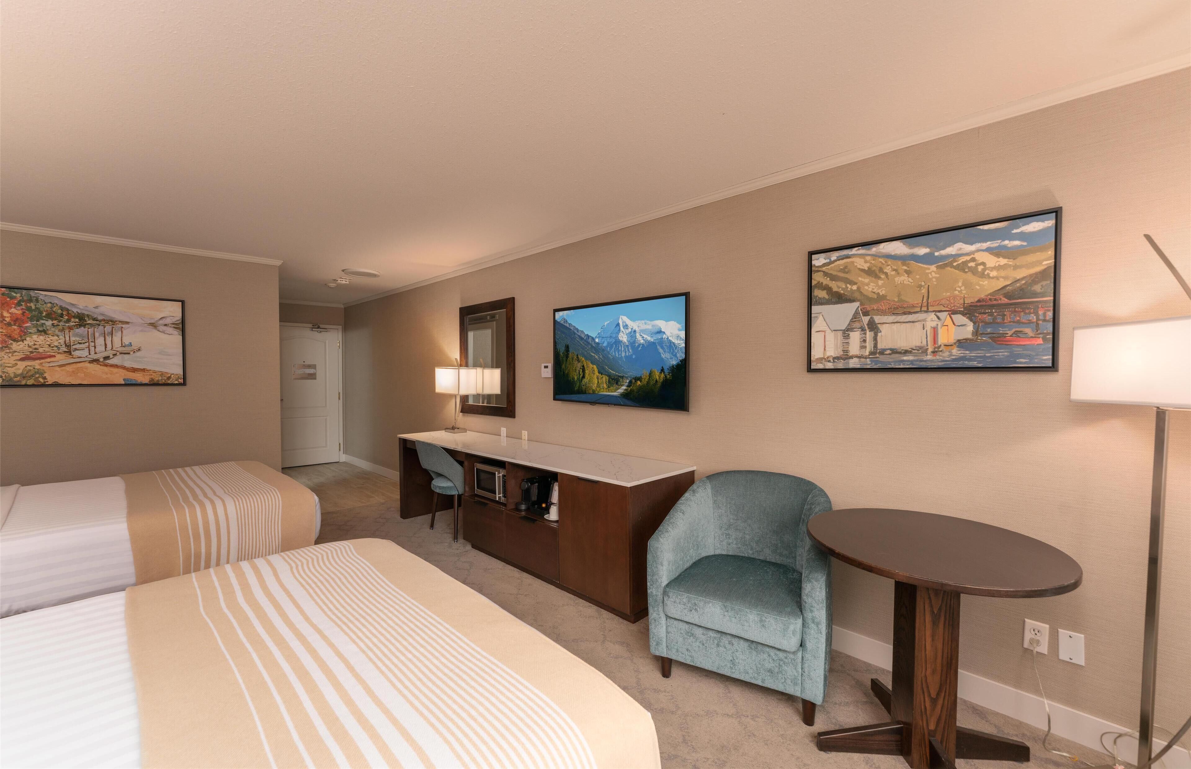 A hotel room with two beds, a desk, a chair, and a small table. There are three paintings on the walls.