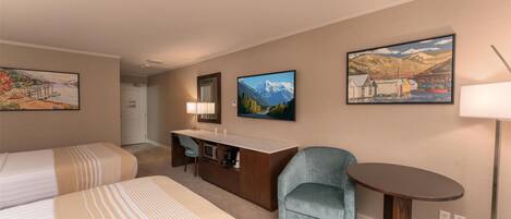 A hotel room with two beds, a desk, a chair, and a small table. There are three paintings on the walls.