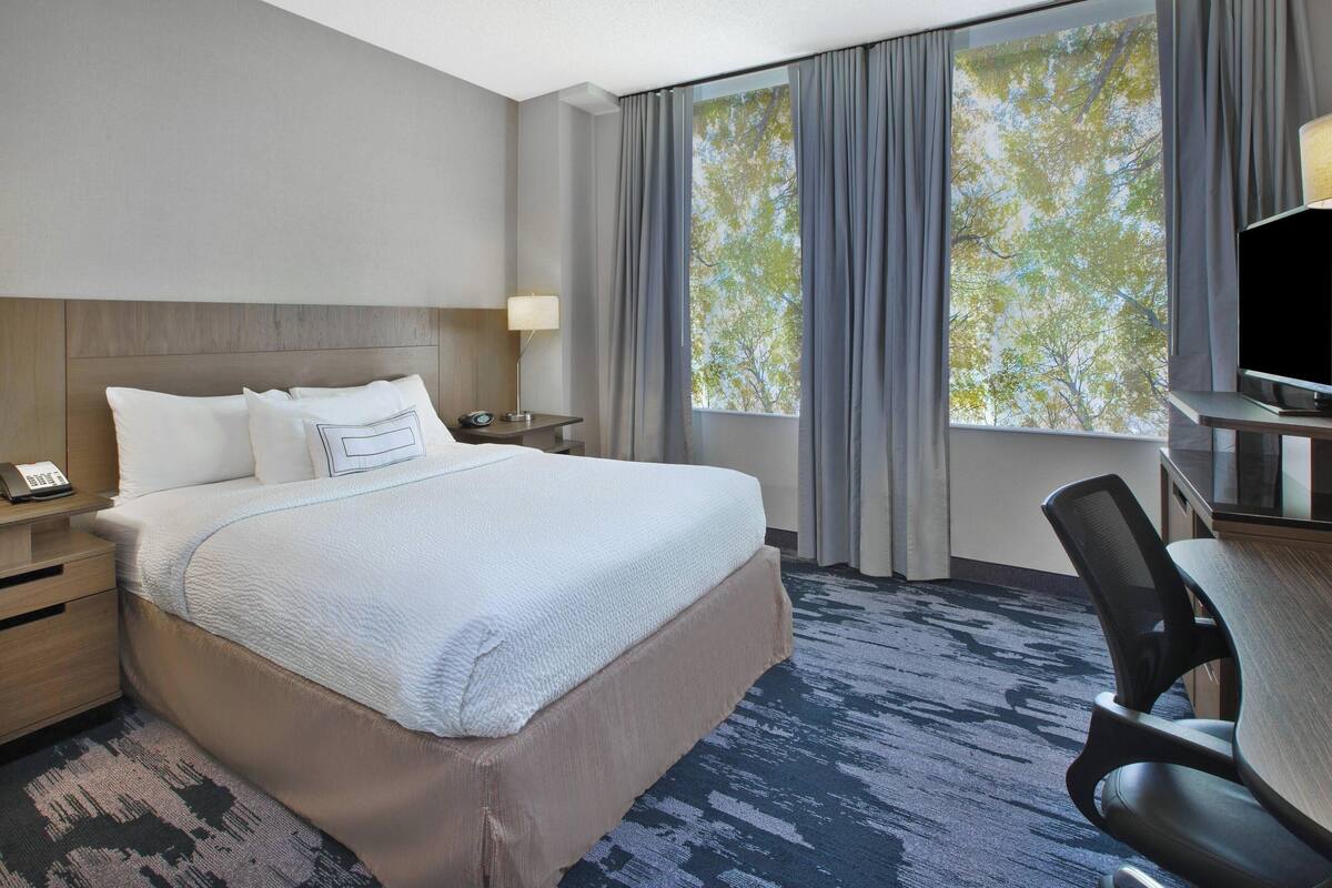 Une chambre d’hôtel avec un grand lit, un bureau avec une chaise, une télévision et une fenêtre avec des rideaux.