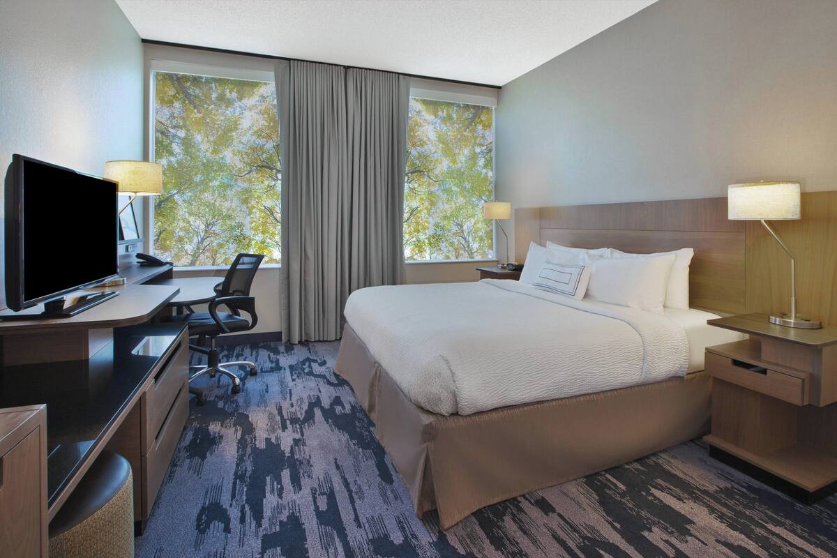 Une chambre d’hôtel équipée d’un lit, d’un bureau, d’une télévision et de deux fenêtres avec des rideaux.