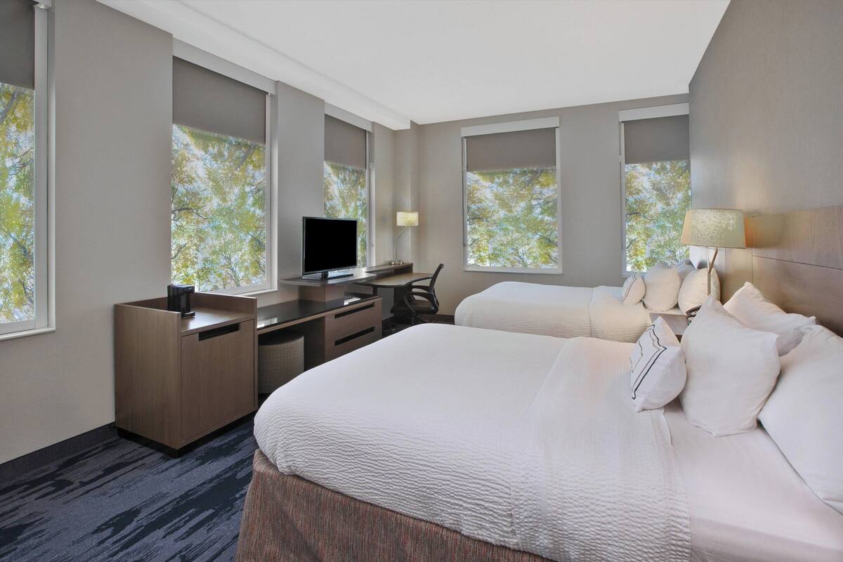 Une chambre d’hôtel équipée d’un lit, d’un bureau, d’une chaise, d’une télévision et de grandes fenêtres donnant sur un paysage arboré.