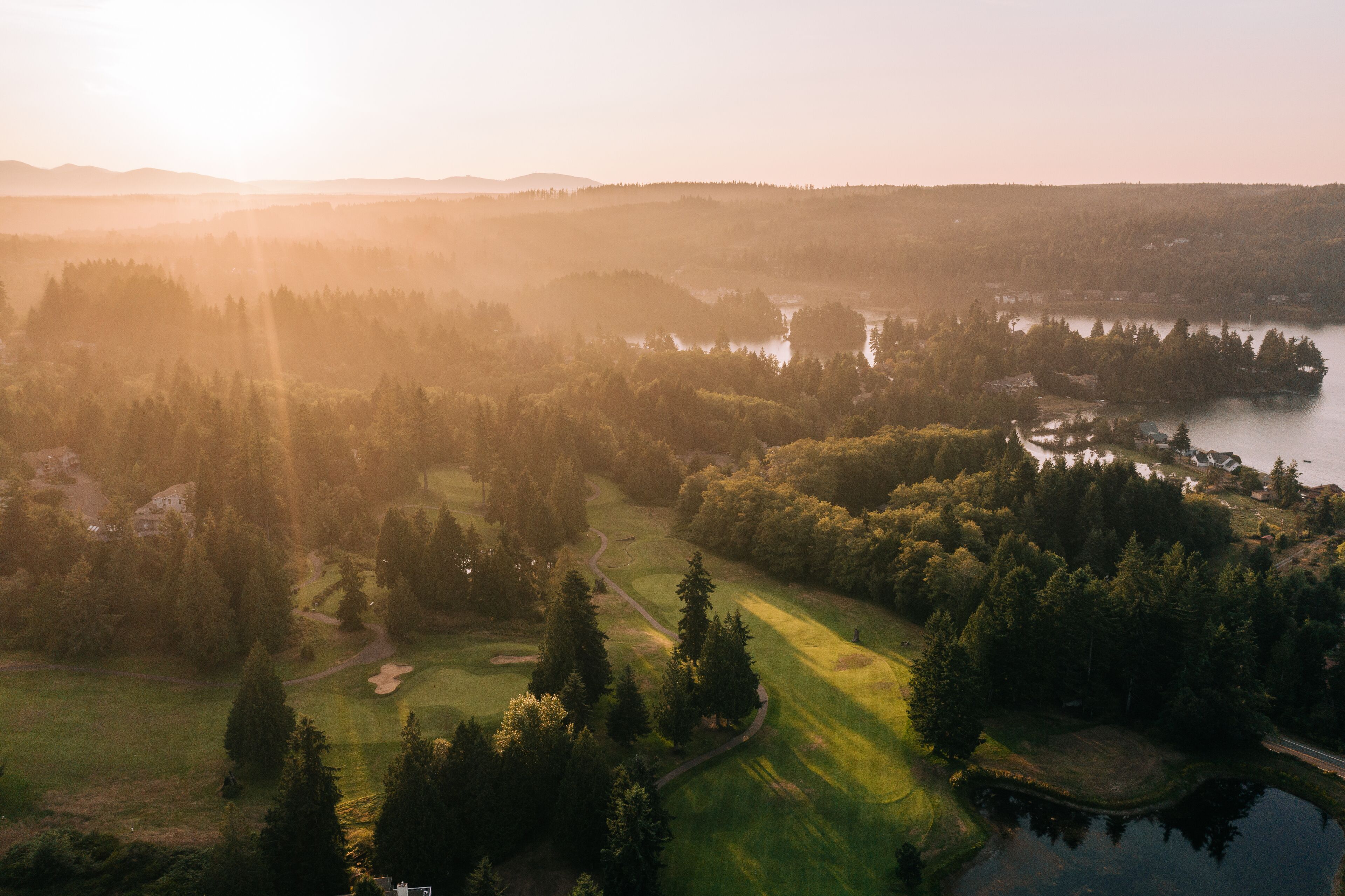 An aerial view of a golf course with a lake, surrounded by dense forests and a few buildings.