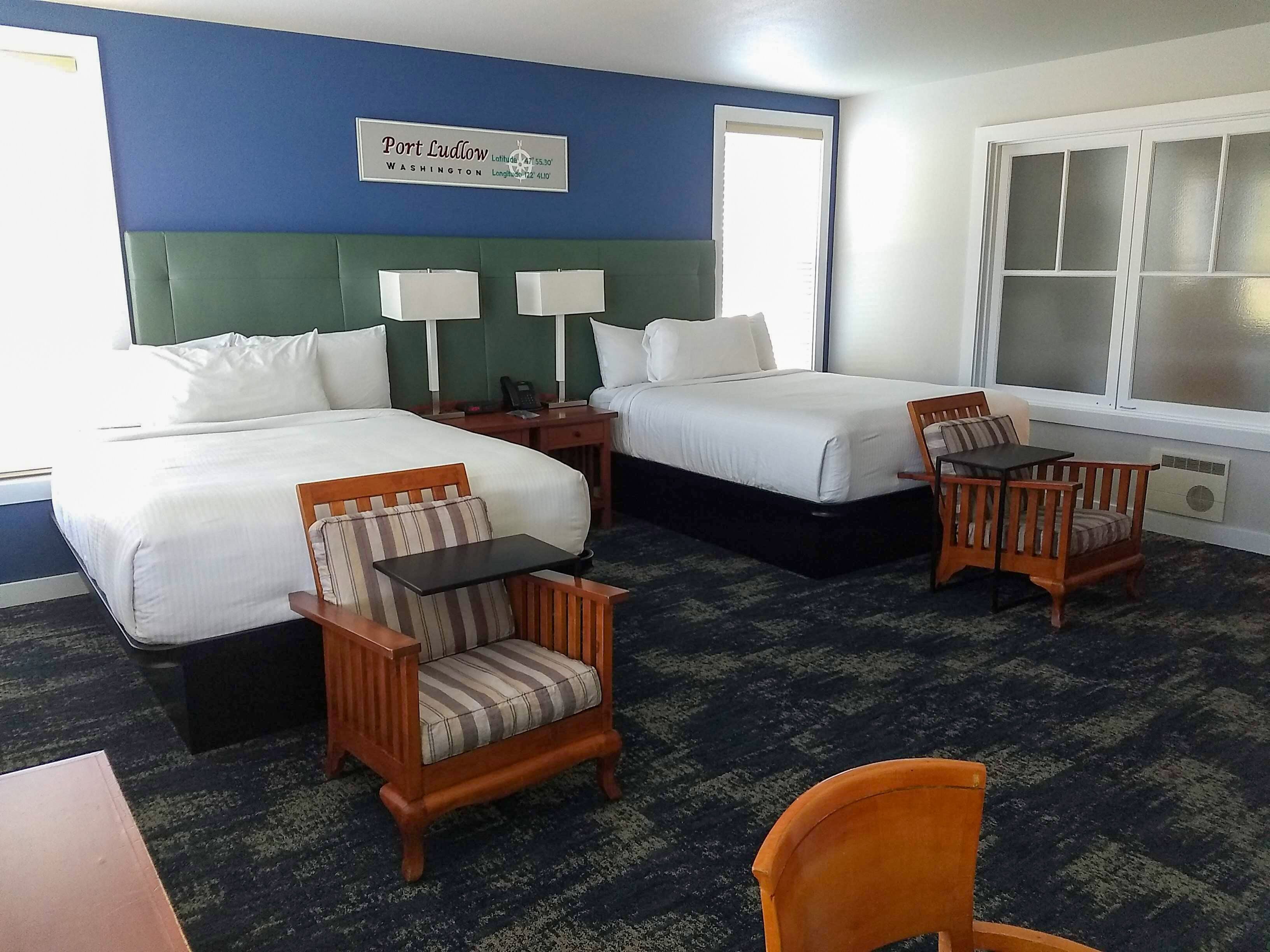 A hotel room with two beds, a small table, two chairs, a nightstand with a lamp, and a sign indicating the location as Port Ludlow, Washington.