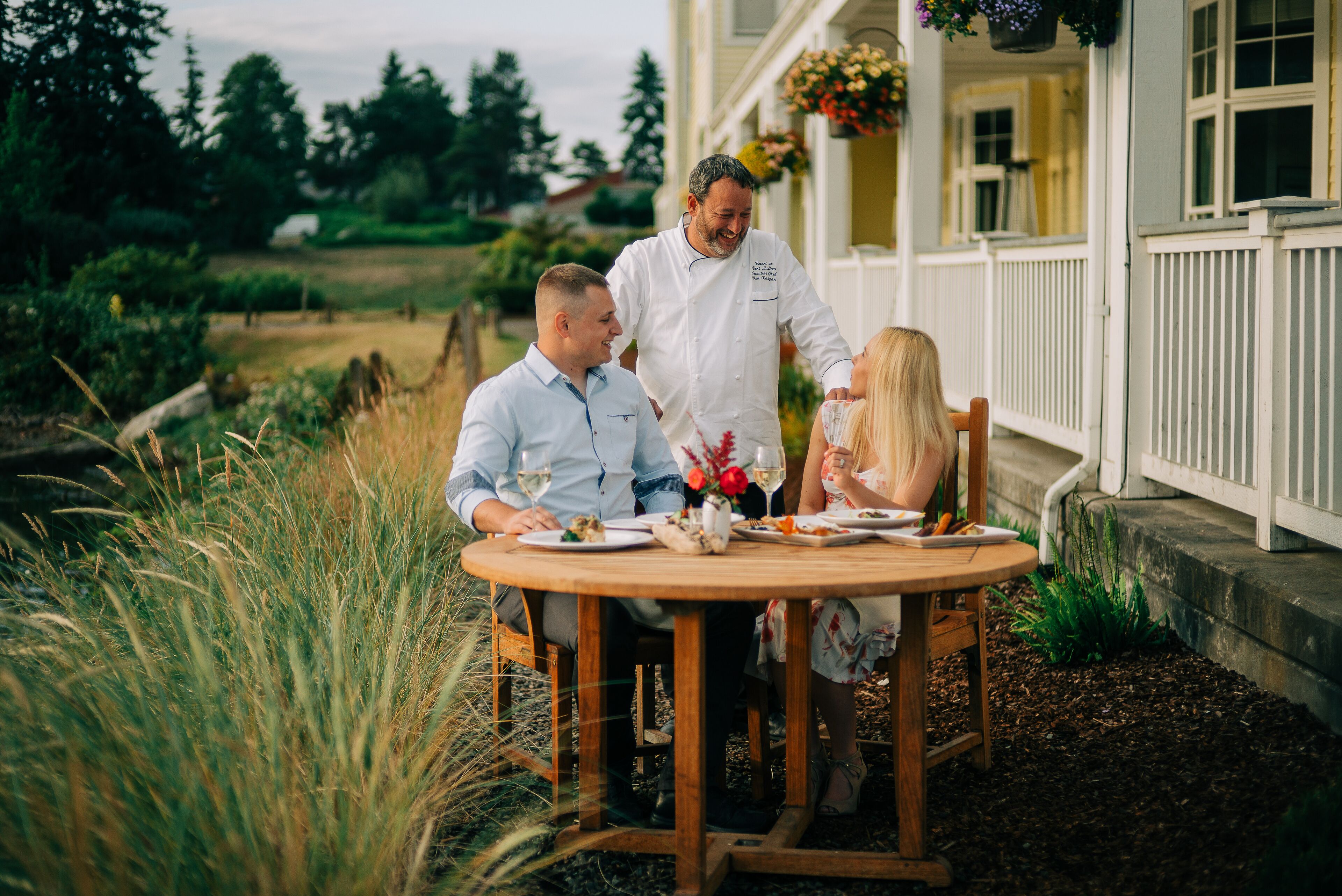 A round wooden table with chairs, a white fence, and a house in the background.