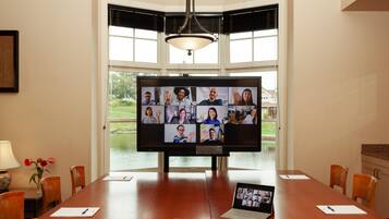 A conference room with a large monitor displaying a video call, a laptop on the table, and a view of a lake through the windows.