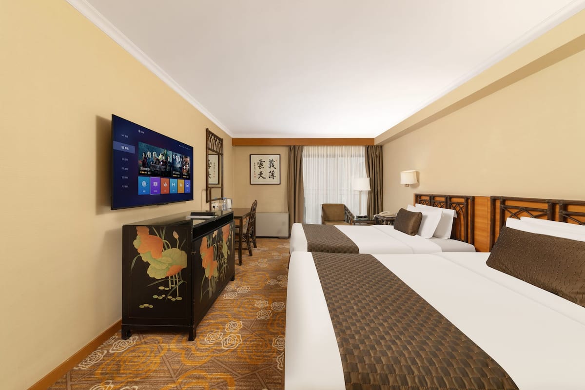 Ein Hotelzimmer mit Flachbildfernseher, Schreibtisch mit Stuhl, zwei Betten, Nachttisch, Lampe und einem gerahmten Bild an der Wand.