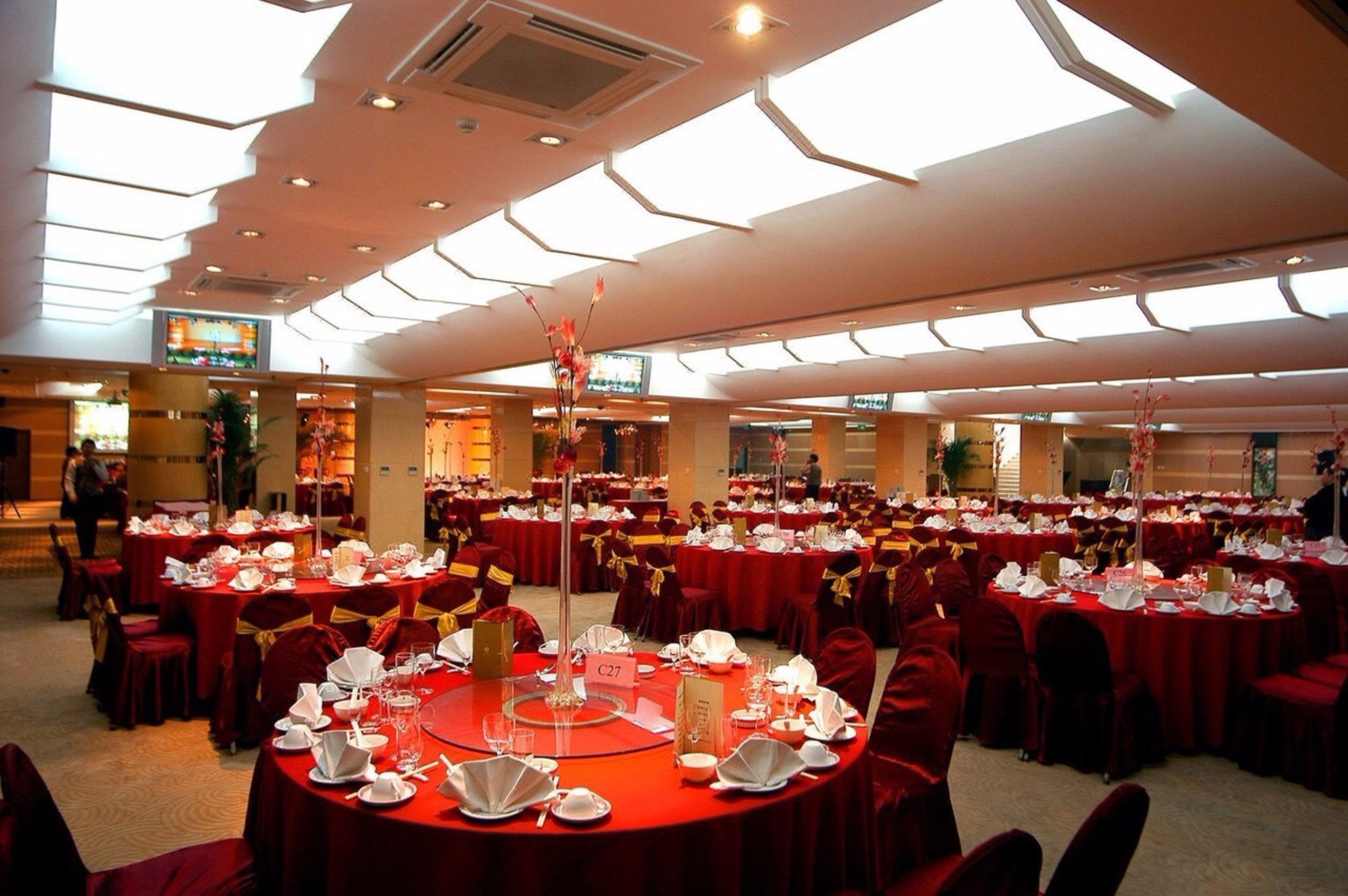 A banquet hall with round tables set for a formal event, featuring a high ceiling with skylights and a central floral arrangement.