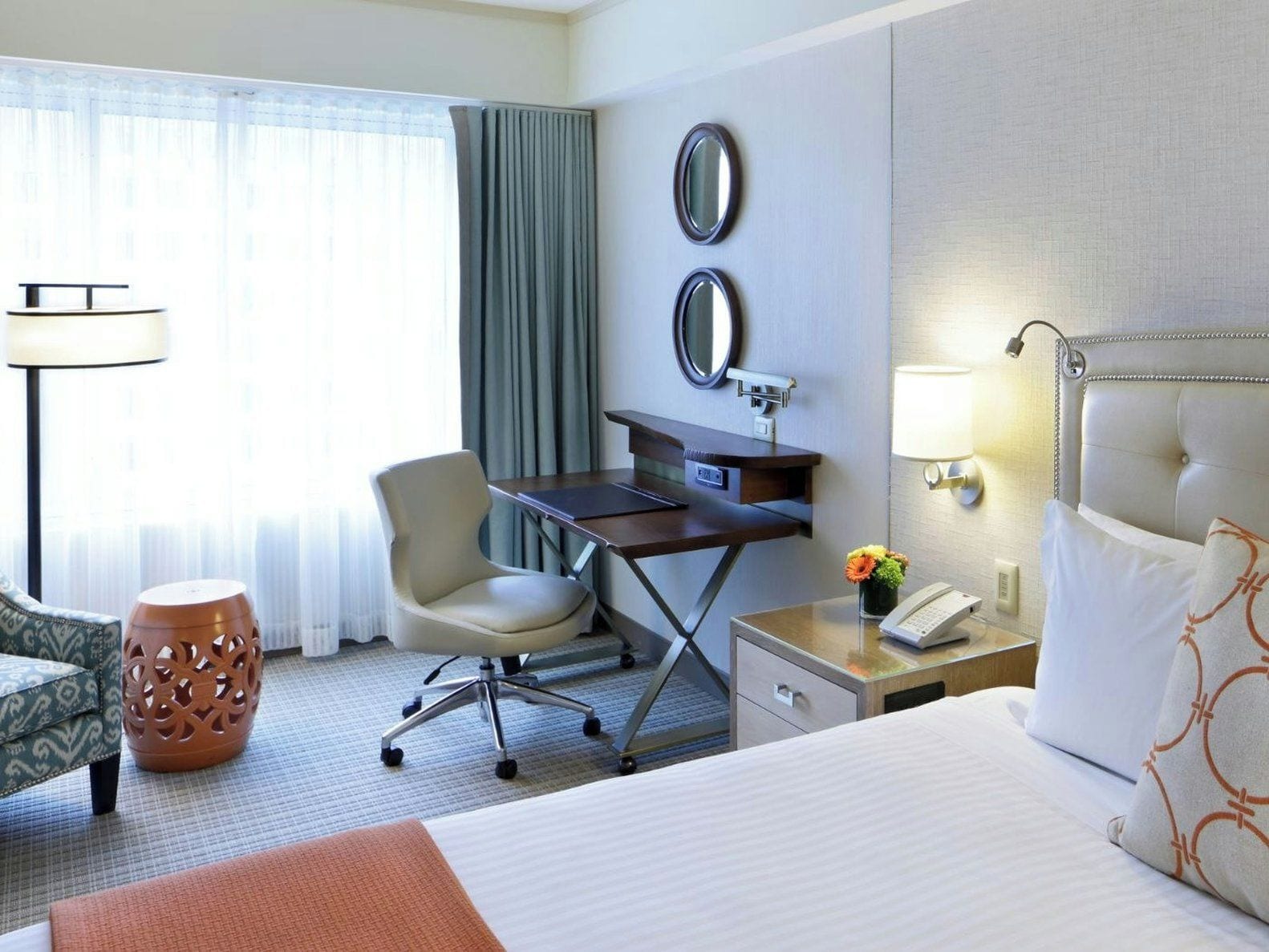 A hotel room with a bed, desk, chair, lamp, and a small table with a phone and flowers.