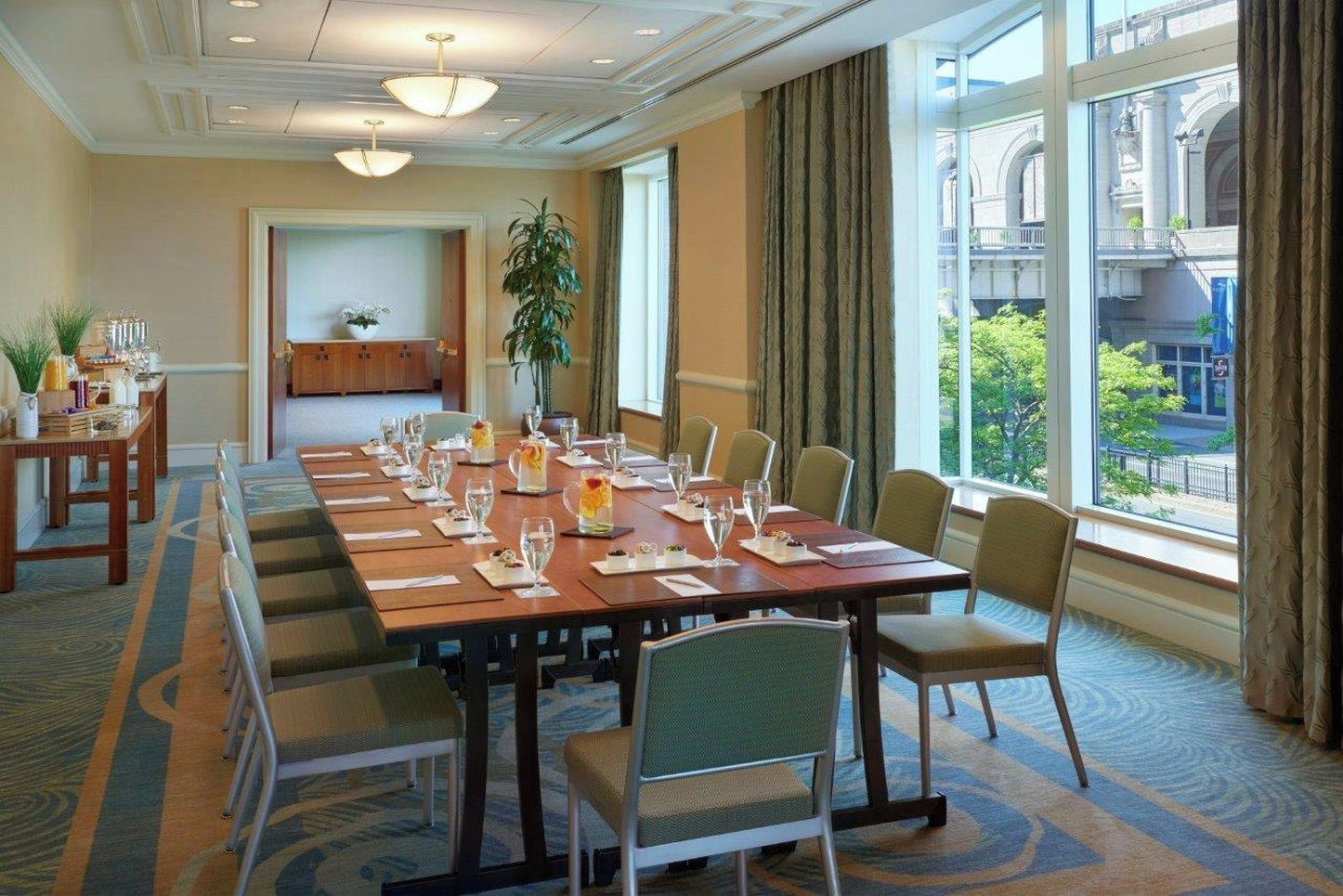 A conference room with a long table set for a meeting, chairs, a plant, and a view of a building through large windows.